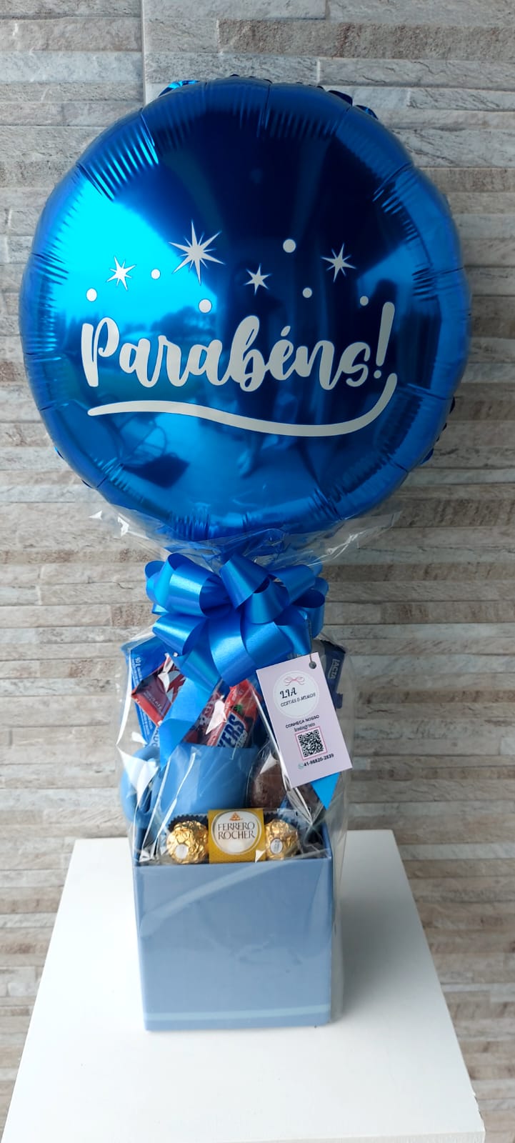 Cesta para presente, com um balão azul, caneca azul e muito chocolate dentro.