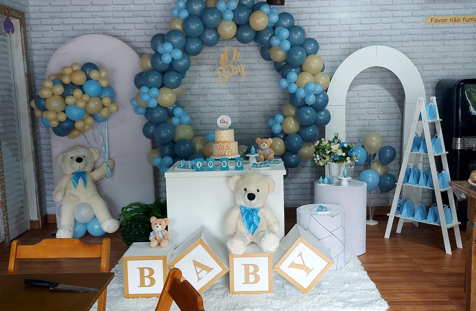 decoração de festa, com ursos de pelúcia, um deles segurando vários balões, de fundo temos uma mesa com enfeite de bolo, e um arco de balão em volta. Os balões são bege escuro e vários tons de azuis